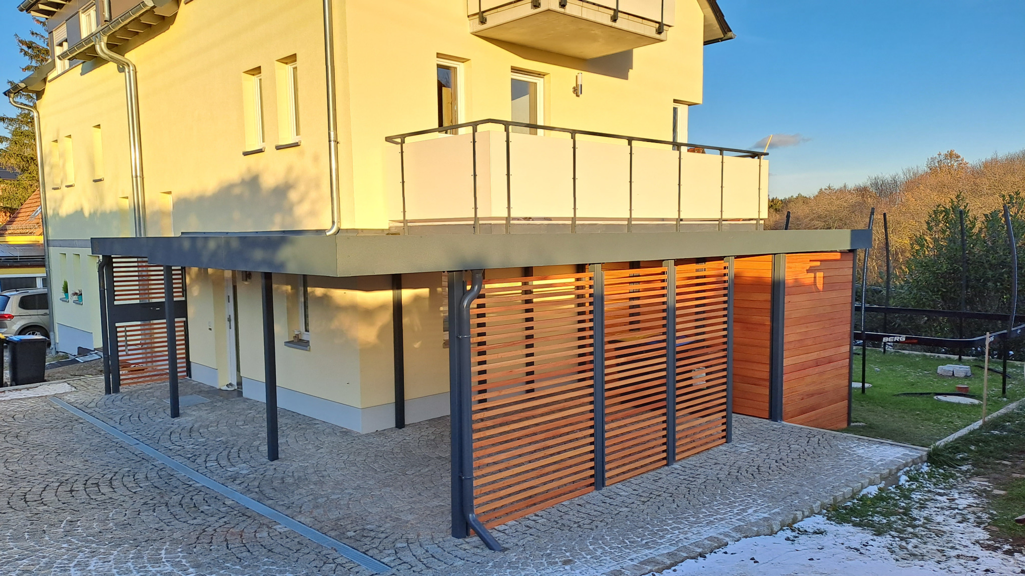 Nowoczesna pergola metalowa z drewnianymi lamelami, przylegająca do jasnego budynku z balkonem. Kostka brukowa na podłożu, widoczne elementy ogrodu.