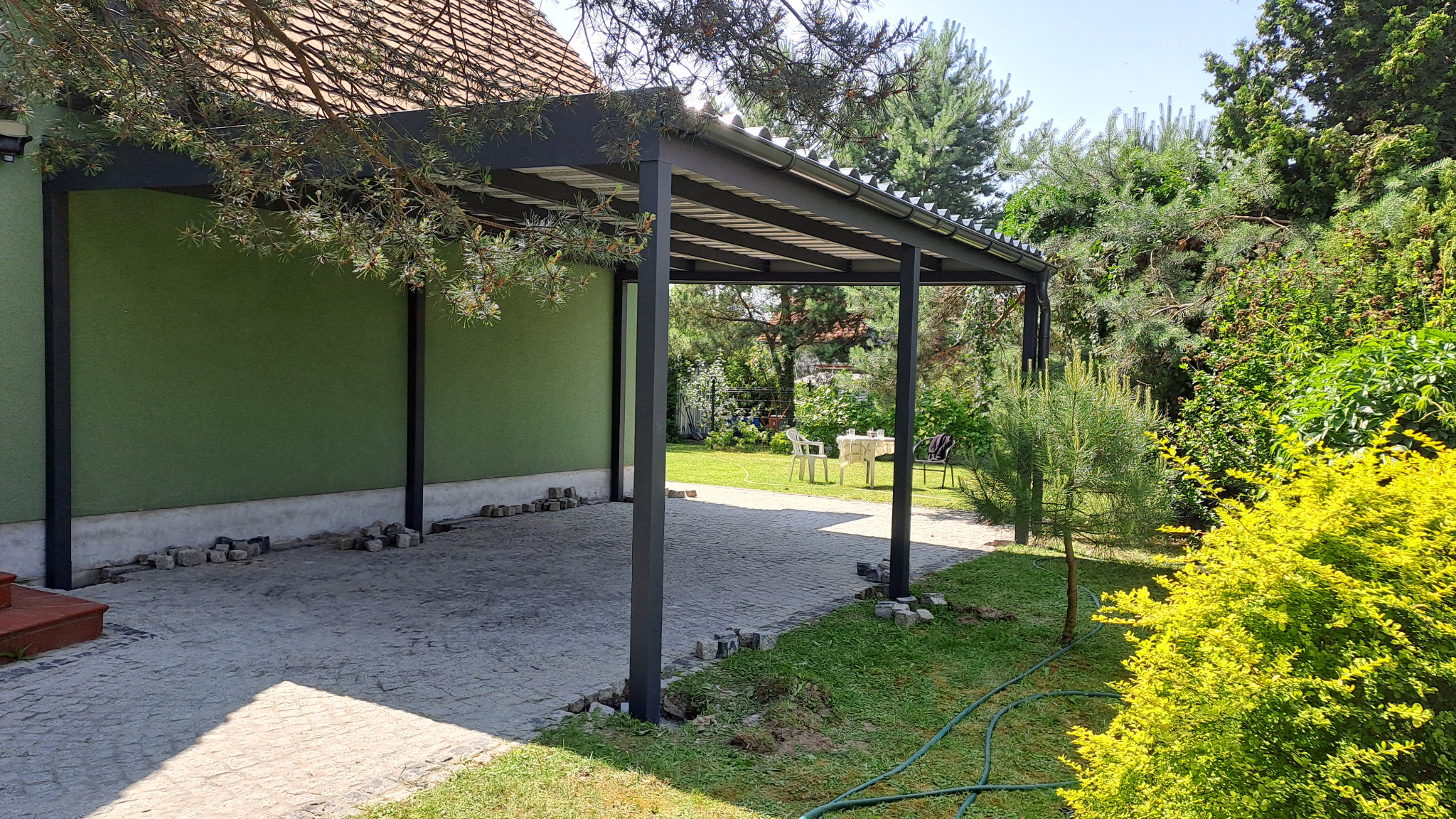 Nowoczesna, grafitowa konstrukcja zadaszenia tarasu przy zielonym domu. Widok na ogród z meblami. Kamienna nawierzchnia i roślinność dookoła.