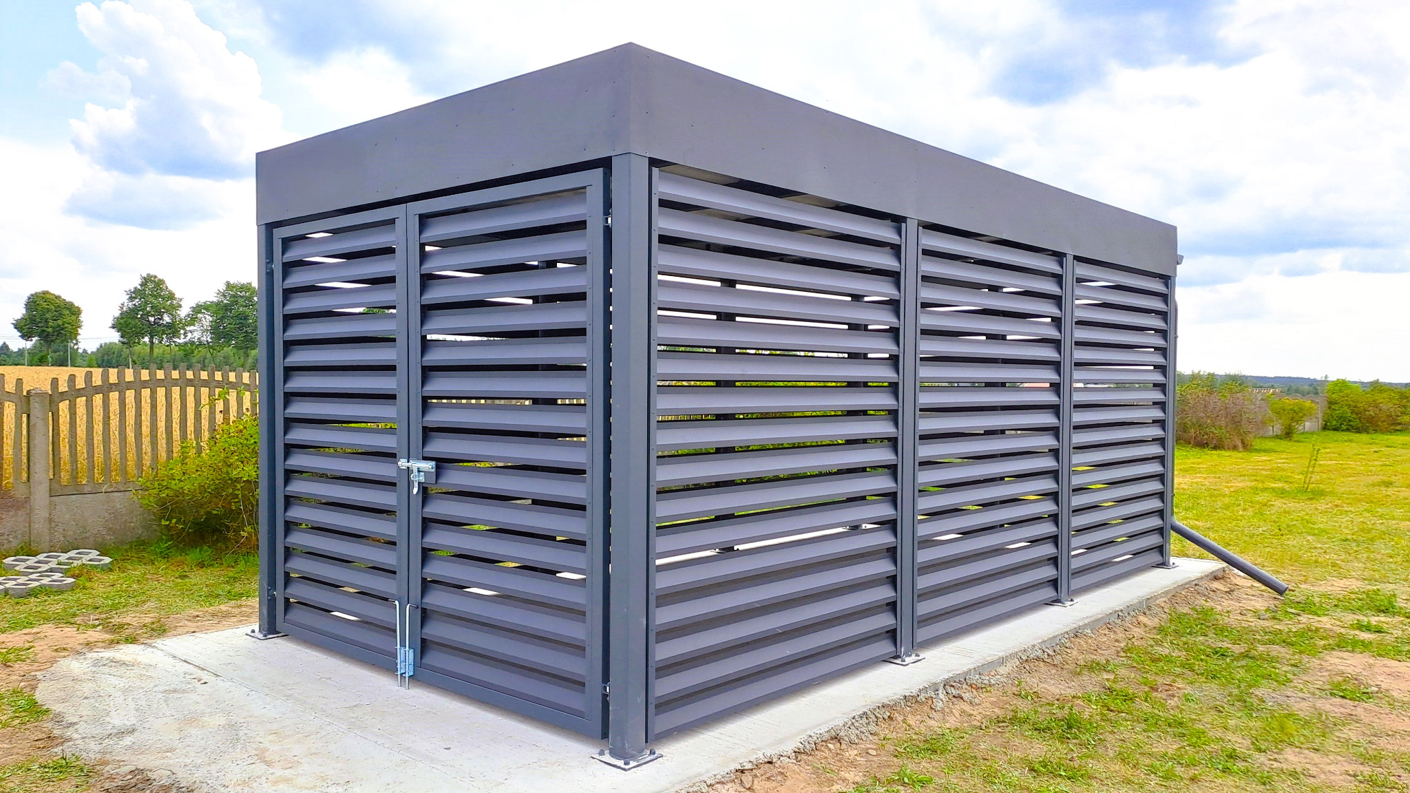 Nowoczesna, grafitowa konstrukcja z poziomych lameli, prawdopodobnie zamykana wiata śmietnikowa, na betonowej podstawie, z rynną odprowadzającą wodę. Tło: zielona trawa.