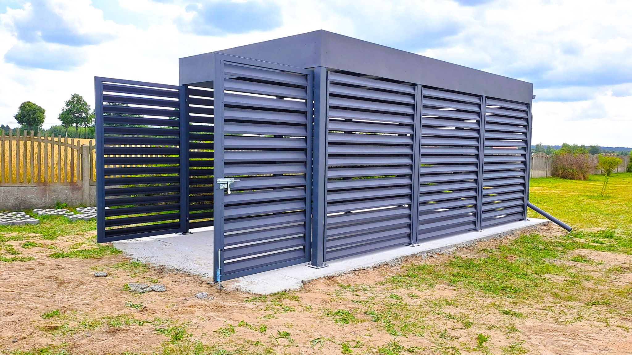 Nowoczesna, szara konstrukcja z metalowych paneli z poziomymi szczelinami, częściowo otwarta, na betonowej podstawie, otoczona trawnikiem i ogrodzeniem.