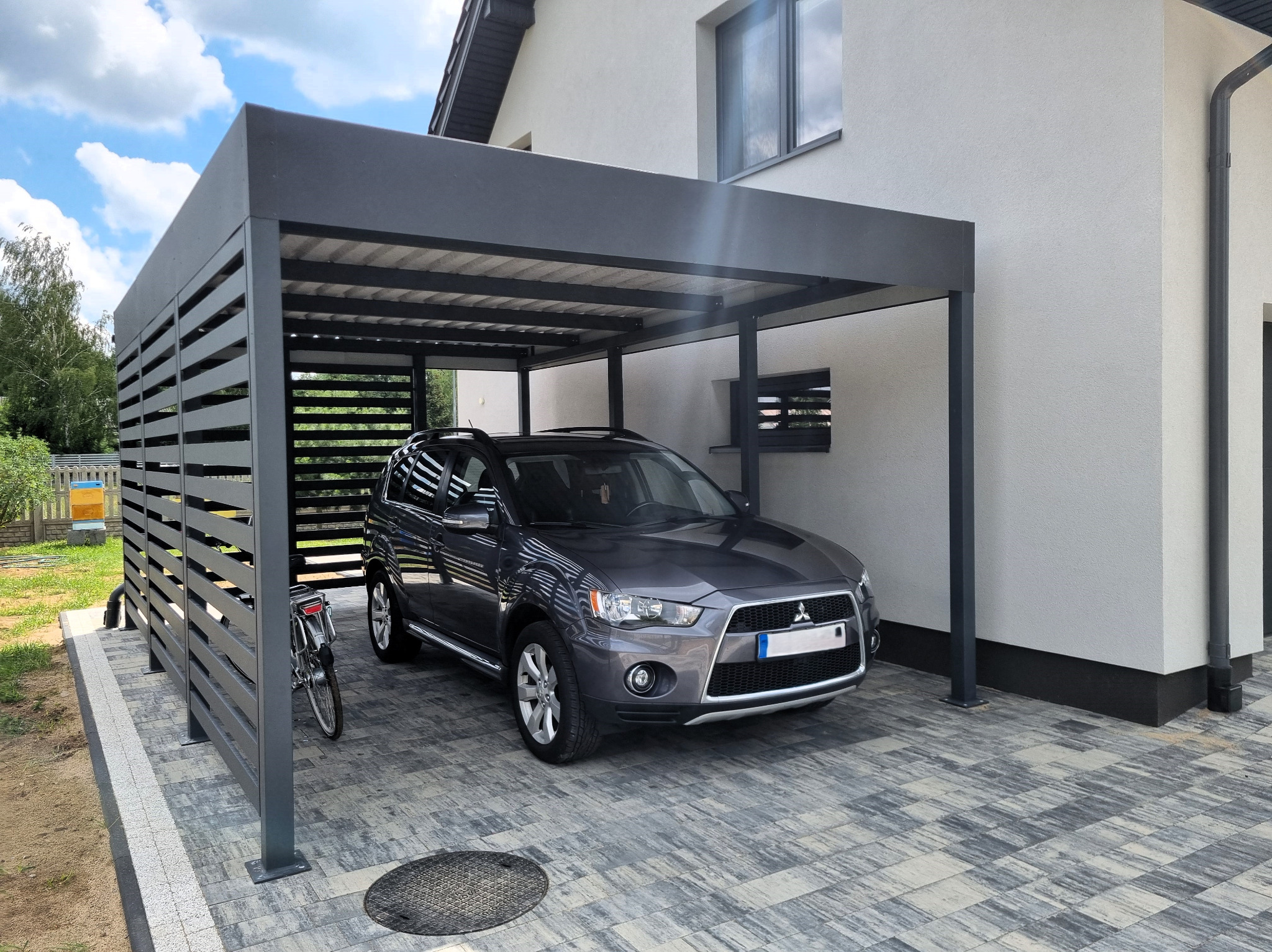 Nowoczesna, grafitowa konstrukcja z metalu, osłaniająca samochód na posesji. Widoczny rower oparty o bok konstrukcji, kostka brukowa i fragment domu.