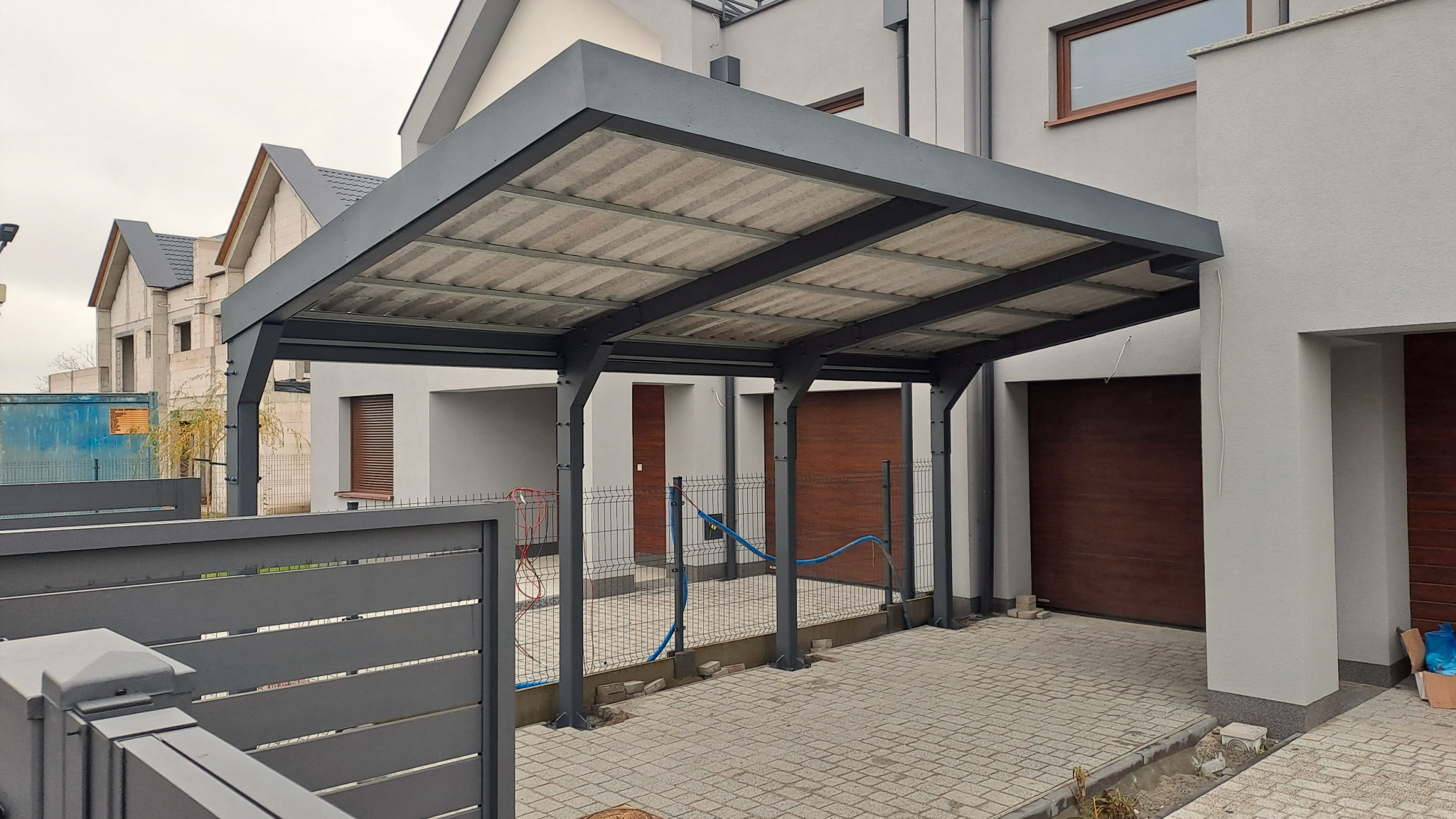 Nowoczesna, grafitowa konstrukcja zadaszenia tarasu z szarej kostki brukowej, widoczna zza ogrodzenia. W tle dom z drewnianą bramą garażową i budynek w trakcie budowy.