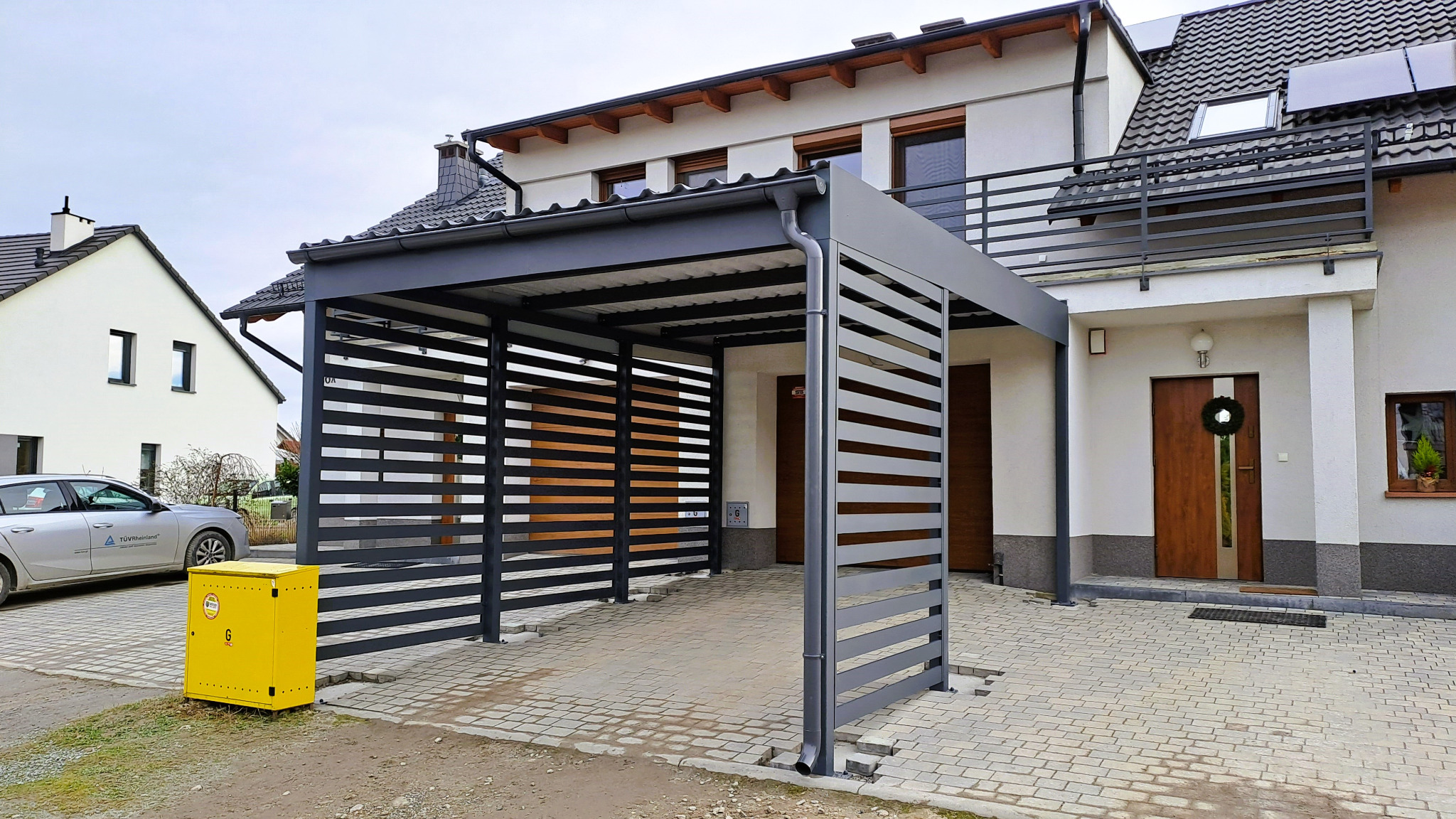 Nowoczesna, szara konstrukcja z poziomych listew, pełniąca funkcję zadaszenia dla samochodu, na tle domu z balkonem i kostki brukowej.