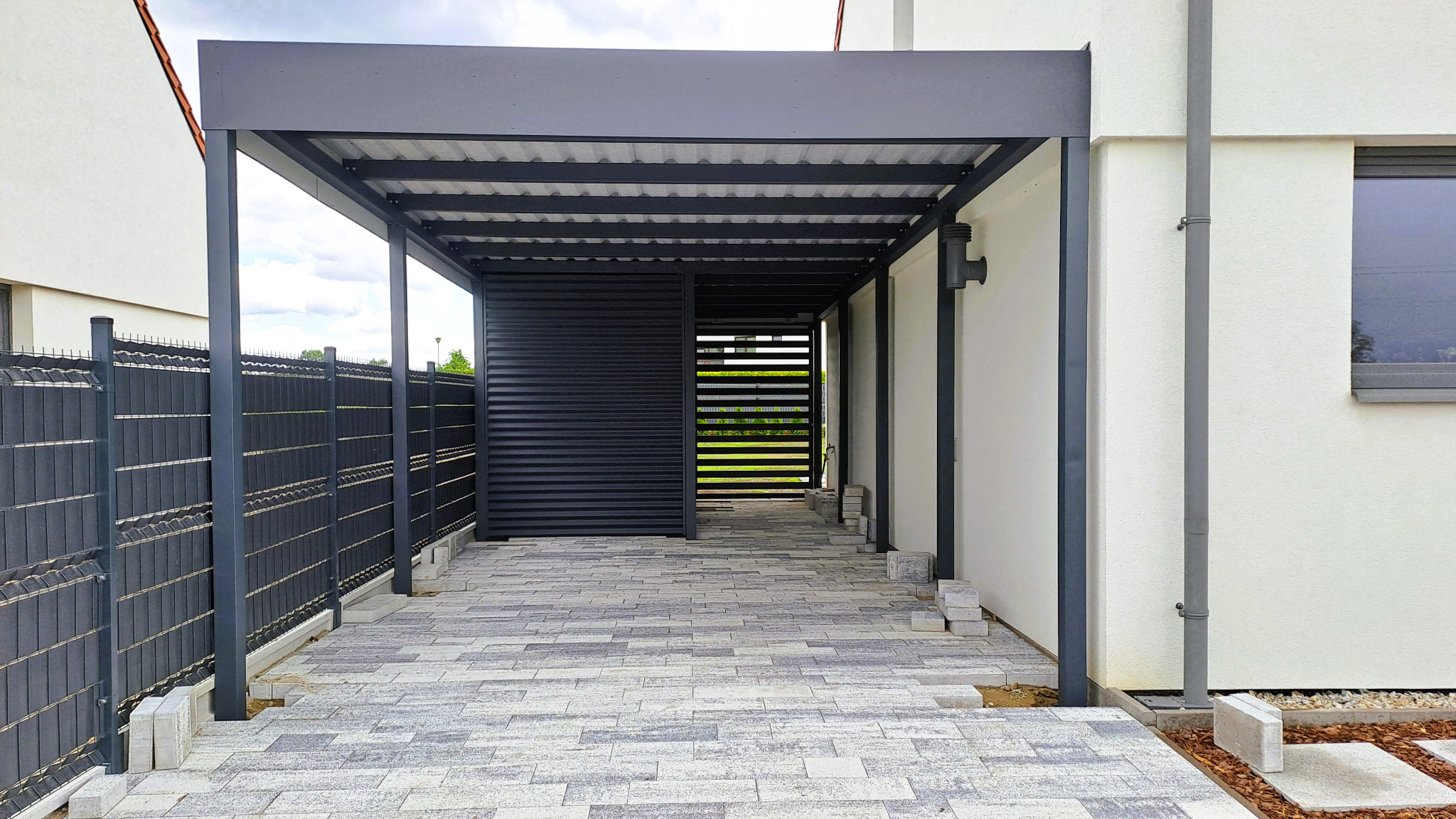 Nowoczesna, grafitowa konstrukcja z metalu, prawdopodobnie z przesuwnym panelem, na tle jasnego domu i ogrodzenia. Widoczna kostka brukowa.