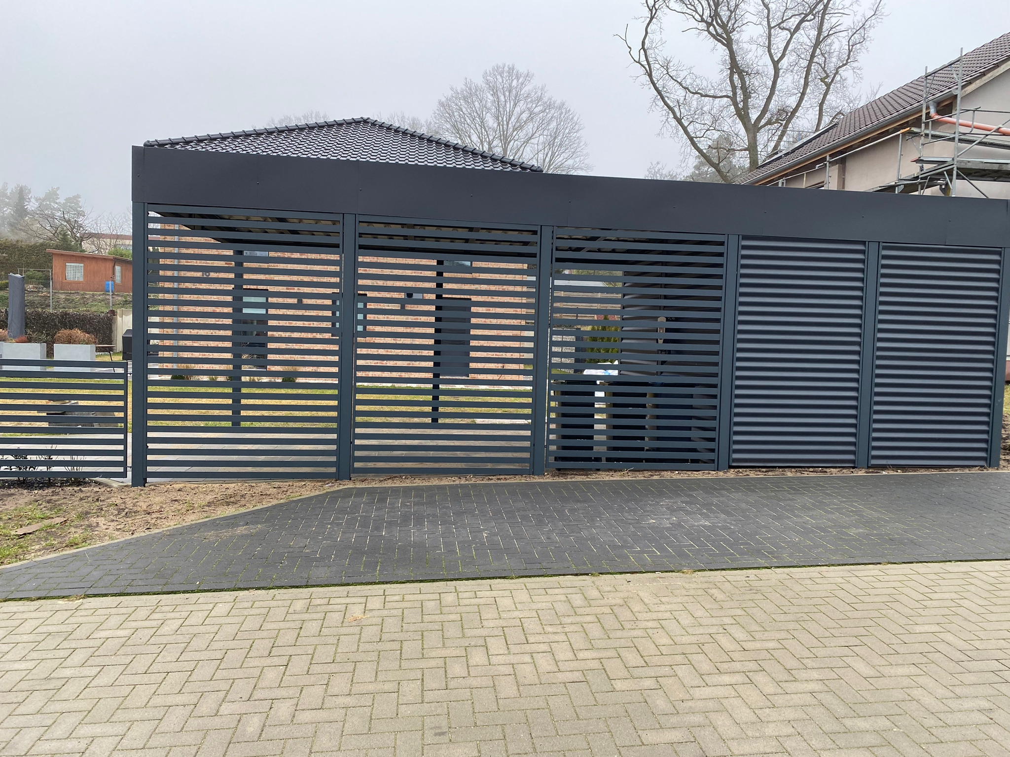 Nowoczesna, grafitowa konstrukcja z poziomych listew, prawdopodobnie zadaszenie tarasu lub wiata, na tle kostki brukowej i zabudowy jednorodzinnej w pochmurny dzień.