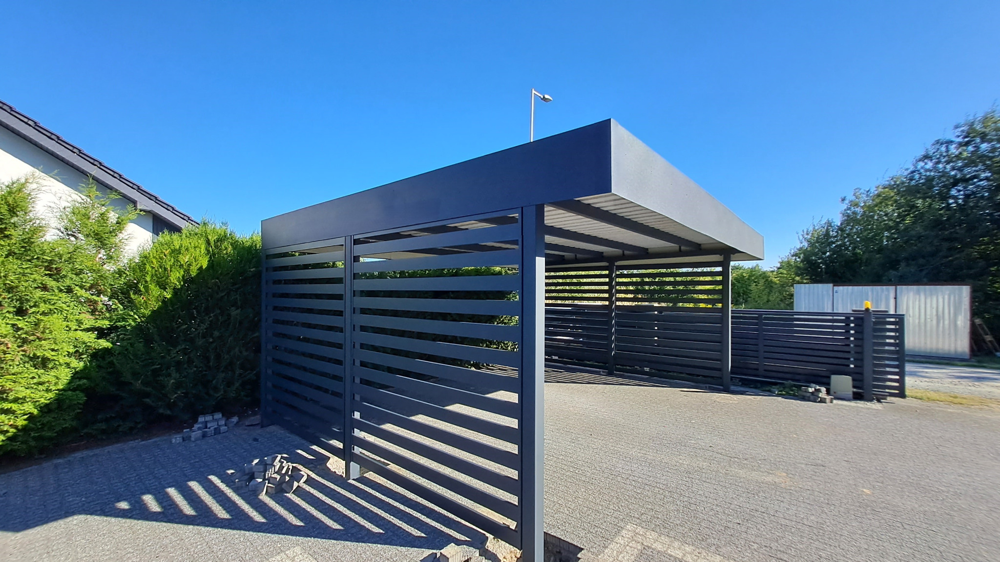 Nowoczesna, metalowa pergola z poziomymi lamelami w kolorze grafitowym, na tle błękitnego nieba i zieleni. Widoczne kostki brukowe i fragment domu.