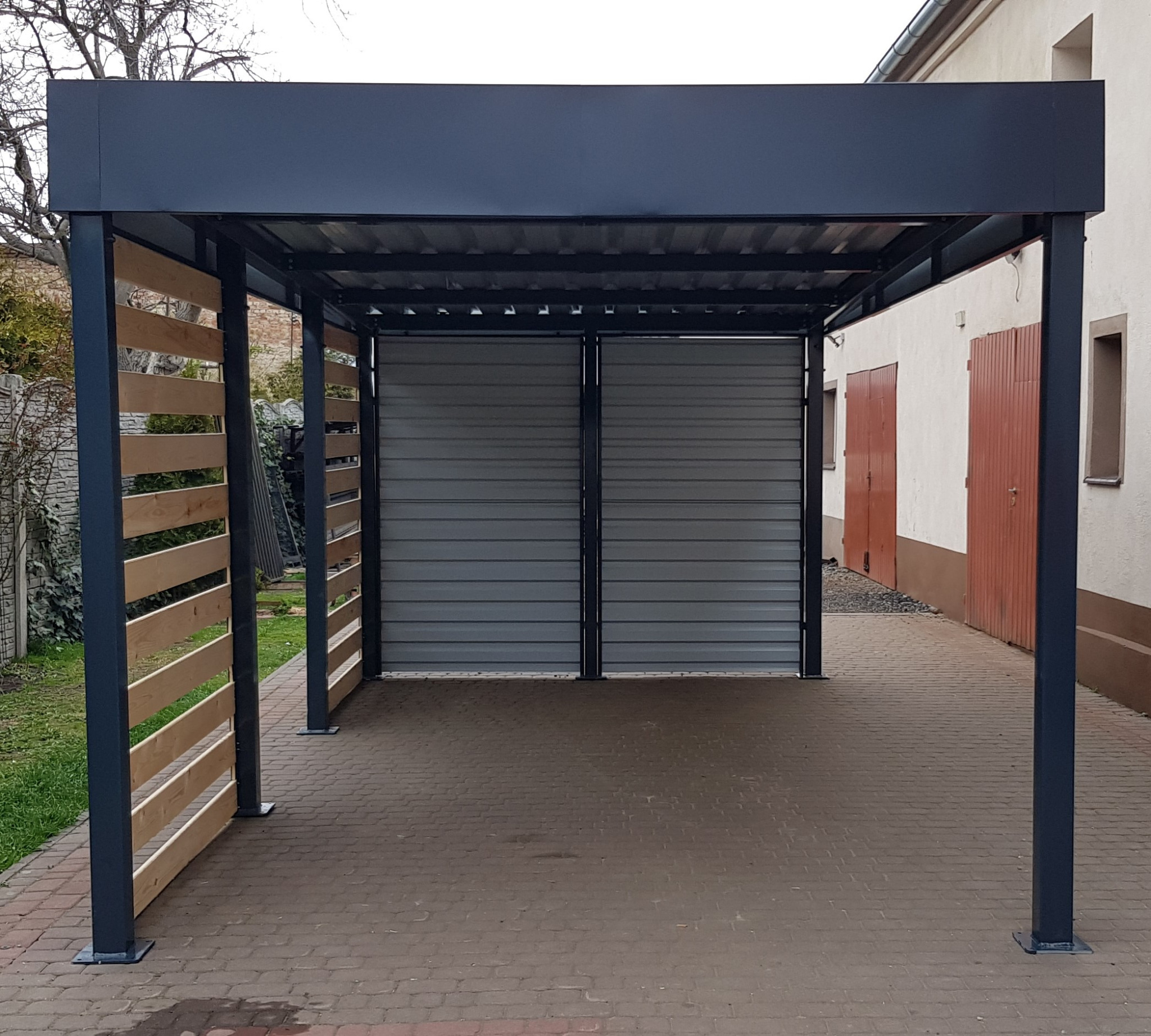 Nowoczesna wiata garażowa z metalową konstrukcją w kolorze grafitowym, częściowo osłonięta drewnianymi panelami, zamykana metalową bramą rolowaną, na podjeździe z kostki brukowej.