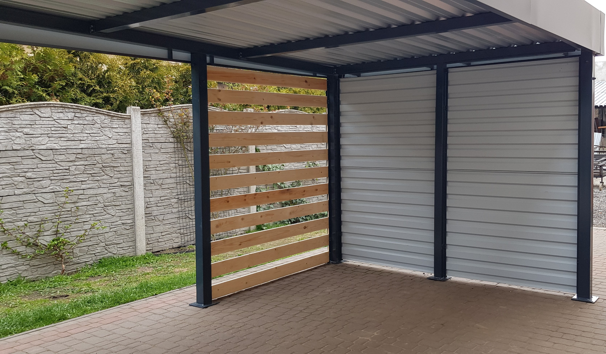 Konstrukcja zadaszenia z metalowym szkieletem i częściowo drewnianą, ażurową ścianą boczną, widoczny fragment posesji z betonowym ogrodzeniem.
