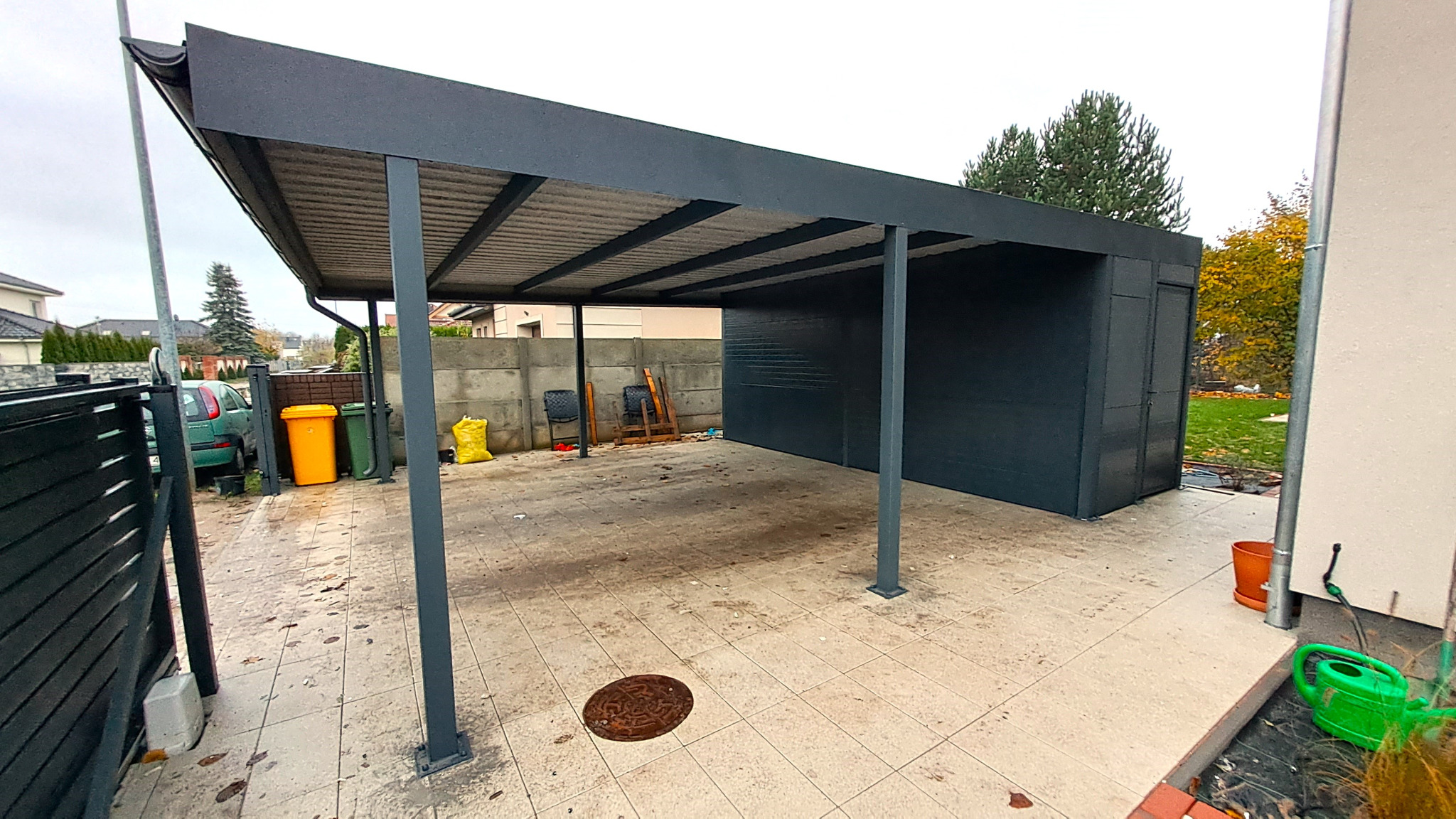 Nowoczesna, grafitowa wiata garażowa z metalową konstrukcją i przylegającym pomieszczeniem gospodarczym, widok z zewnątrz na betonowej kostce, z elementami otoczenia jak kosze na śmieci i fragment...