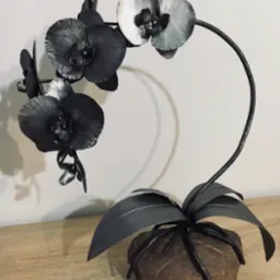 Metalowa rzeźba przedstawiająca orchideę z liśćmi i kwiatami, osadzona na kamieniu, stojąca na drewnianym blacie.