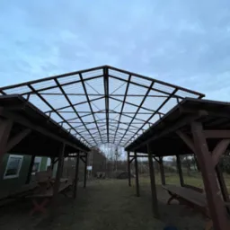 Zadaszenie dwóch drewnianych altan połączonych metalową konstrukcją z przezroczystym dachem, widok z dołu na tle pochmurnego nieba.