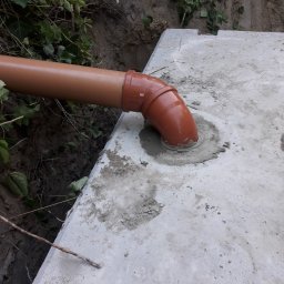 Zakład usługowy Lewek Grzegorz - Brązowa rura kanalizacyjna wchodzi pod kątem w betonową płytę, uszczelniona zaprawą cementową, widoczna krawędź wykopu i zieleń.