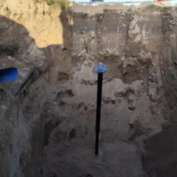 Głęboki wykop w Rokietnicy z wystającą czarną rurą, zabezpieczoną niebieskim przewodem na górze, ściany wykopu z wyraźnymi warstwami ziemi, w tle siatka ogrodzeniowa i fragment samochodu.