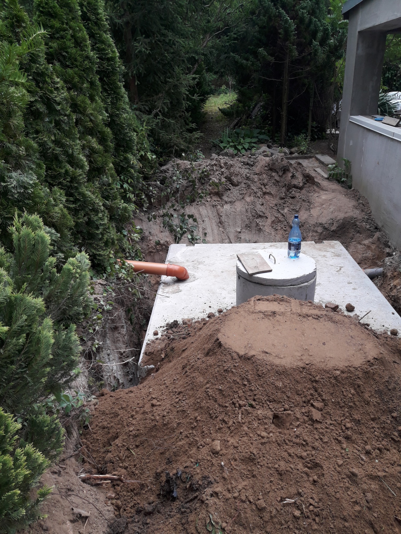Betonowy zbiornik, częściowo zakopany w ziemi, z widocznym wlotem rury kanalizacyjnej, obok pryzma ziemi i butelka wody na górze, w tle zieleń.
