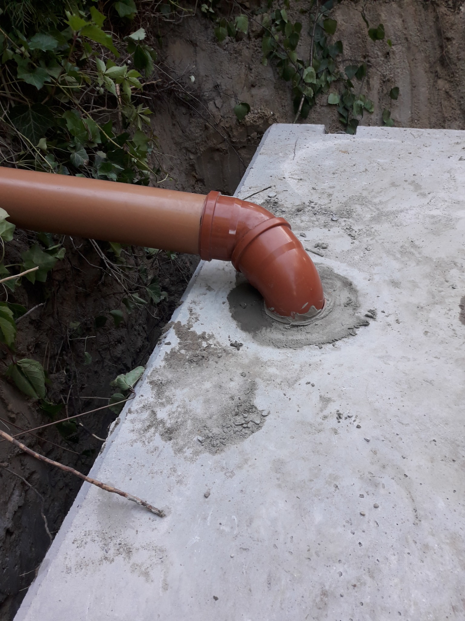 Brązowa rura kanalizacyjna wchodzi pod kątem w betonową płytę, uszczelniona zaprawą cementową, widoczna krawędź wykopu i zieleń.