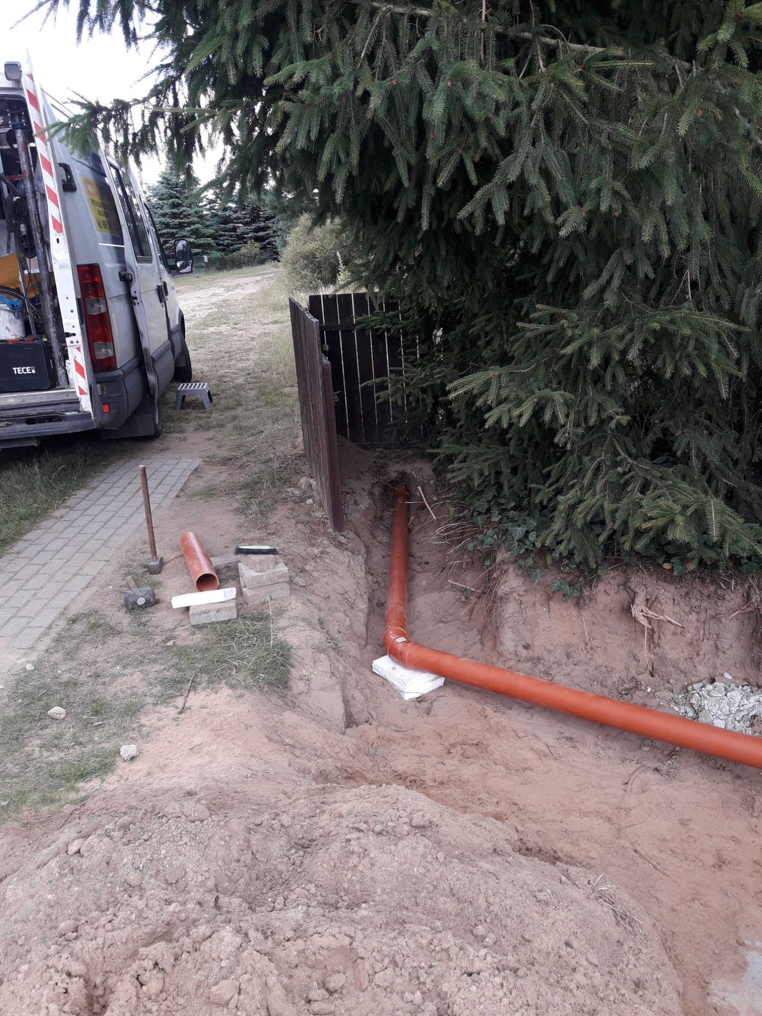 Wykopany rów z ułożonym pomarańczowym rurociągiem kanalizacyjnym, widoczny samochód dostawczy z otwartymi drzwiami, narzędzia i materiały budowlane, teren częściowo zarośnięty trawą, w tle drzewa...