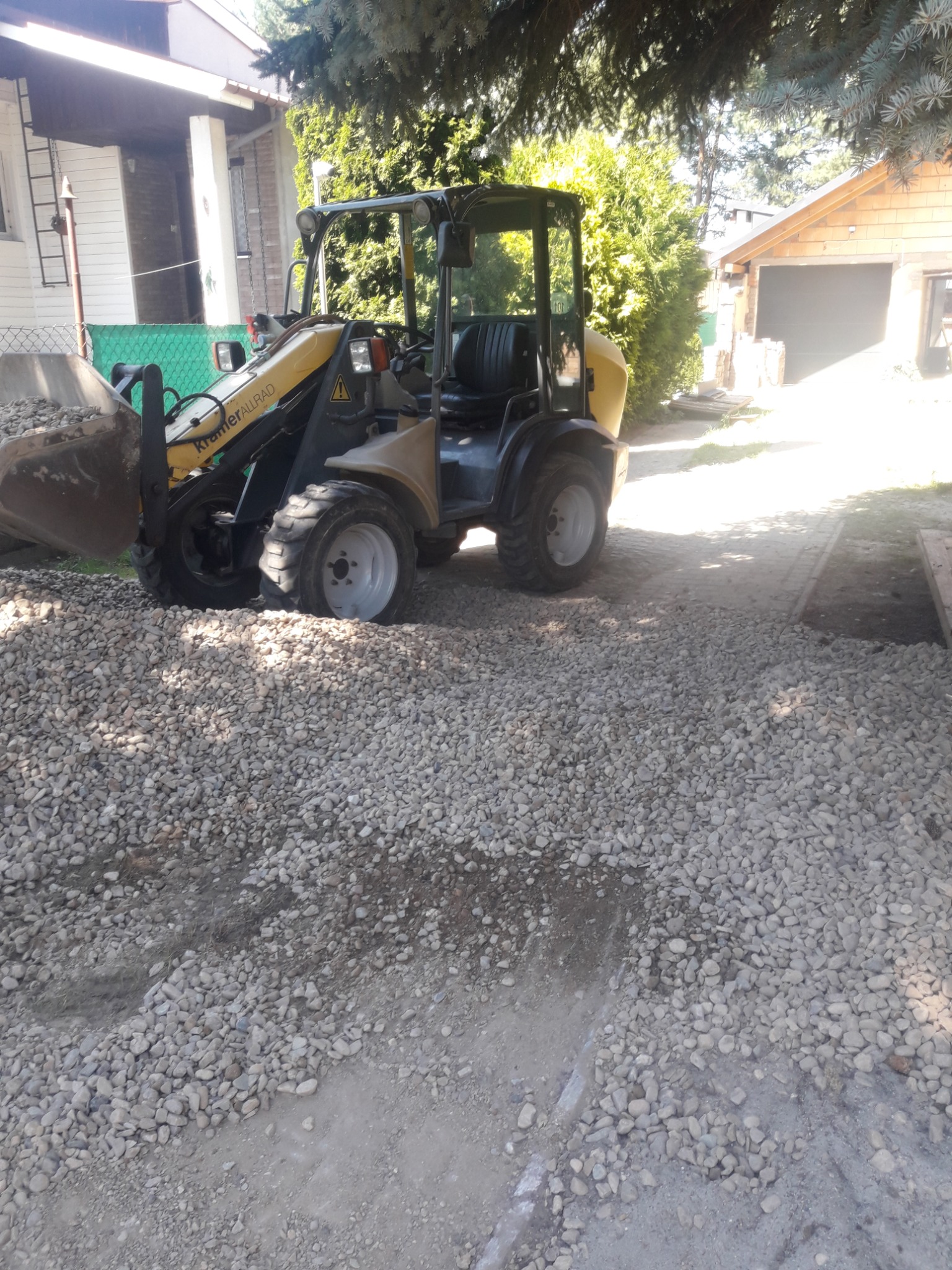 Żółta ładowarka Kramer Allrad częściowo zasypana żwirem na terenie budowy, z widocznym domem i garażem w tle.