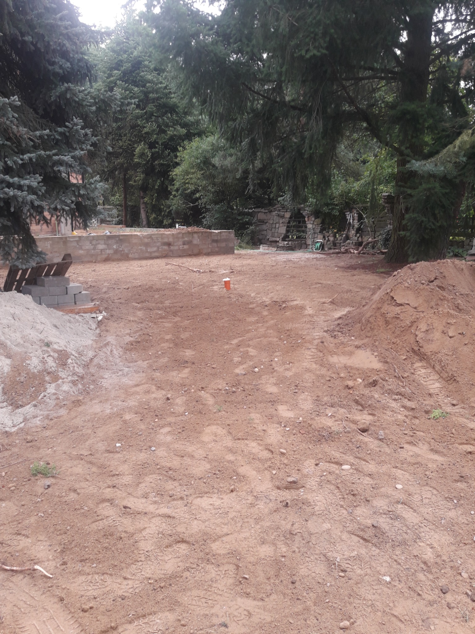 Fundamenty budynku w trakcie budowy, widoczne bloczki betonowe, wykopana ziemia i otaczająca zieleń drzew.