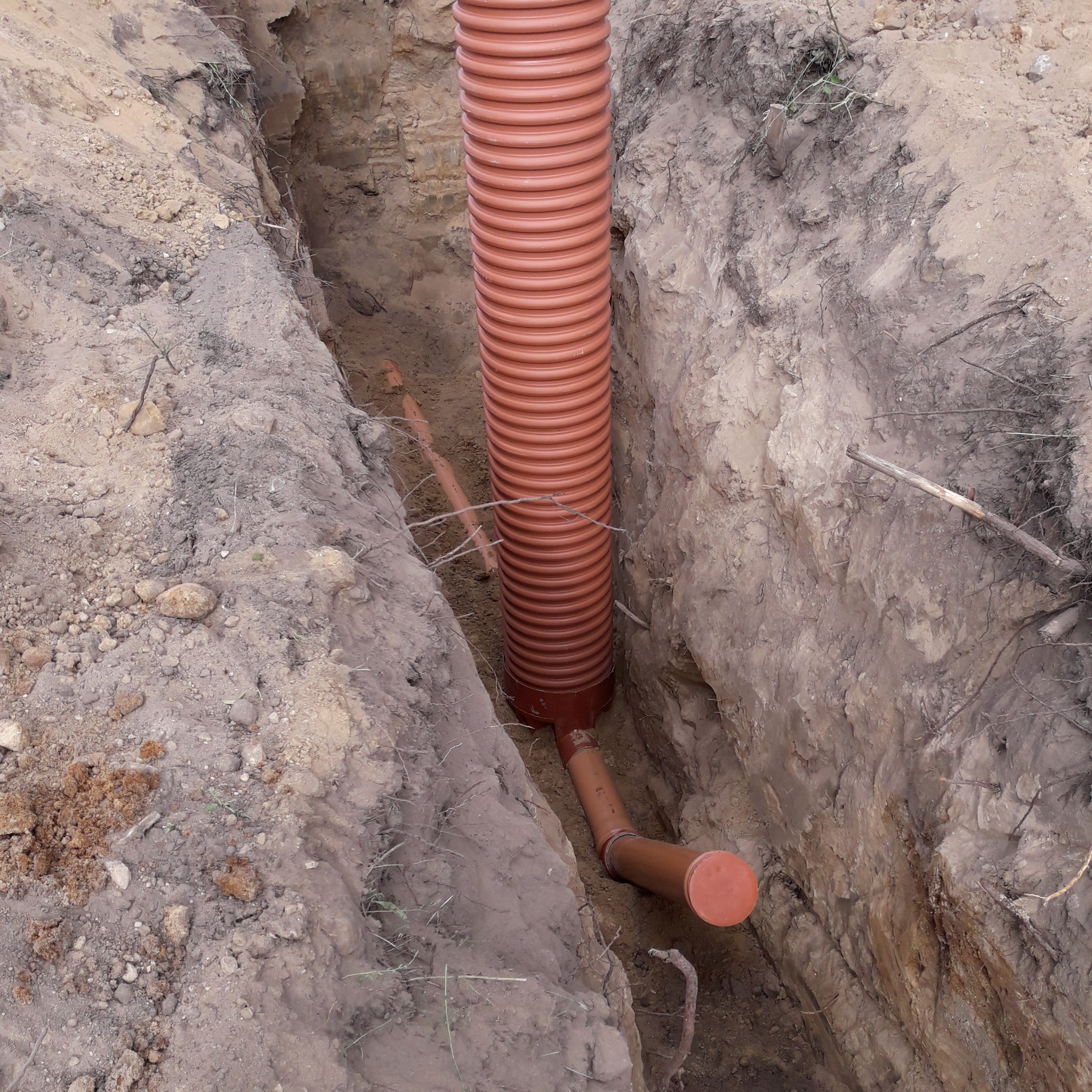 Wykop w ziemi z pionowo umieszczoną, karbowaną rurą z tworzywa sztucznego w kolorze terakoty, połączoną z poziomym odcinkiem rury, z widoczną ziemią i korzeniami.