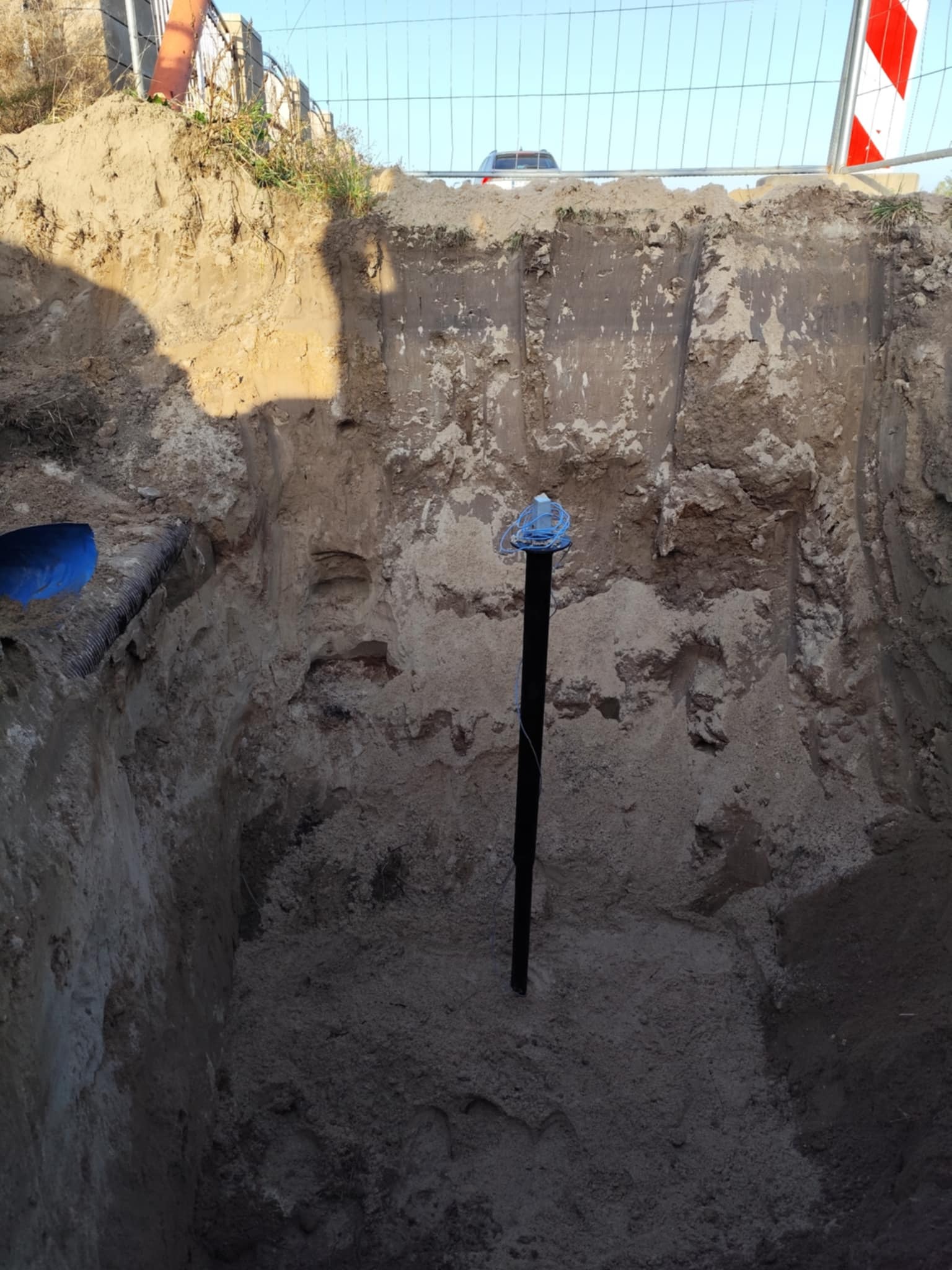 Głęboki wykop w Rokietnicy z wystającą czarną rurą, zabezpieczoną niebieskim przewodem na górze, ściany wykopu z wyraźnymi warstwami ziemi, w tle siatka ogrodzeniowa i fragment samochodu.
