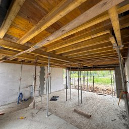 Sharim Invest Sp. Z o. o. - Wnętrze budynku w trakcie budowy; widoczne drewniane szalunki stropu podparte metalowymi stemplami, ściany z białego bloczka, betonowe filary, drabina i fragment instalacji hydraulicznej.