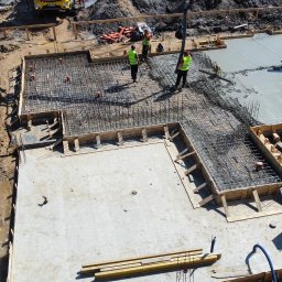 Sharim Invest Sp. Z o. o. - Zalewanie betonem zbrojonej płyty fundamentowej domu jednorodzinnego, widok z góry, pracownicy w kamizelkach odblaskowych nadzorują proces, widoczny szalunek i pompa do betonu.