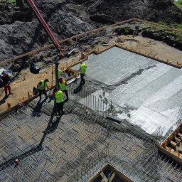 Sharim Invest Sp. Z o. o. - Wylewanie betonu na przygotowane zbrojenie fundamentów budynku, widok z góry, pracownicy w kamizelkach odblaskowych obsługują pompę do betonu.