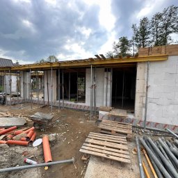 Sharim Invest Sp. Z o. o. - Budowa domu jednorodzinnego w stanie surowym otwartym: ściany z betonu komórkowego, podpory stropowe, widoczne materiały budowlane na placu budowy, pochmurne niebo.