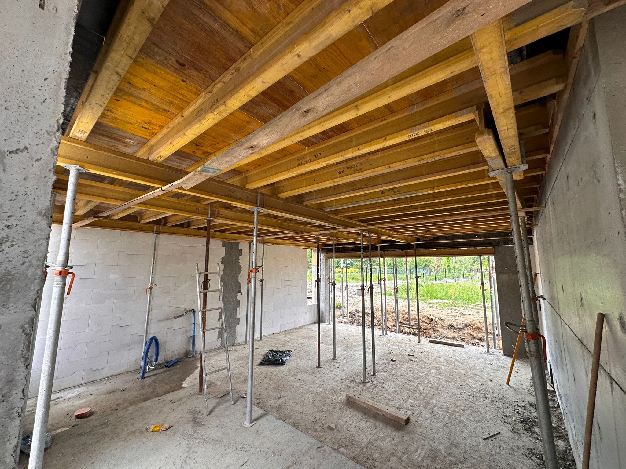 Wnętrze budynku w trakcie budowy; widoczne drewniane szalunki stropu podparte metalowymi stemplami, ściany z białego bloczka, betonowe filary, drabina i fragment instalacji hydraulicznej.