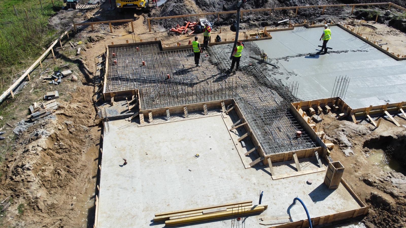 Zalewanie betonem zbrojonej płyty fundamentowej domu jednorodzinnego, widok z góry, pracownicy w kamizelkach odblaskowych nadzorują proces, widoczny szalunek i pompa do betonu.