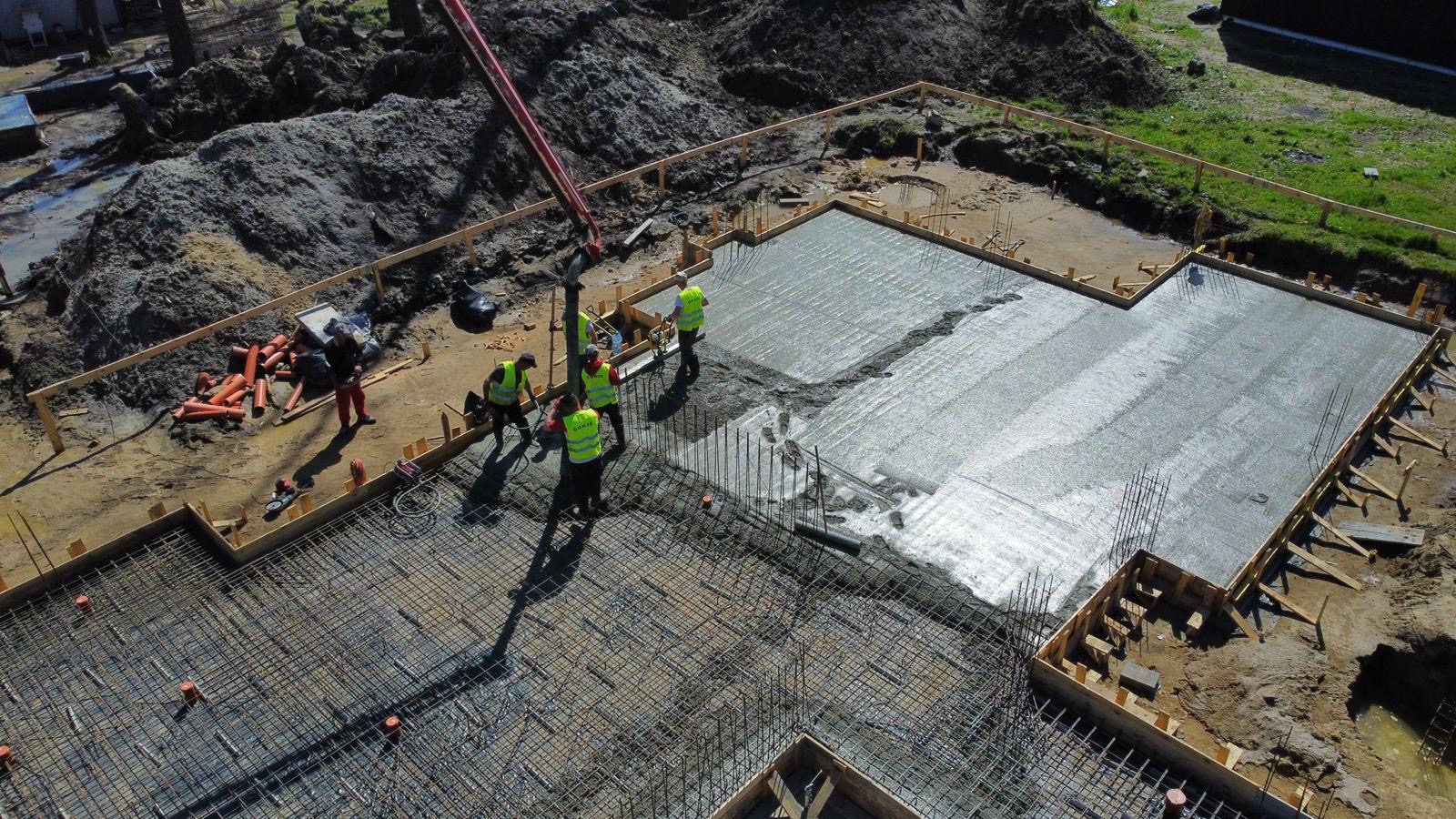 Wylewanie betonu na przygotowane zbrojenie fundamentów budynku, widok z góry, pracownicy w kamizelkach odblaskowych obsługują pompę do betonu.