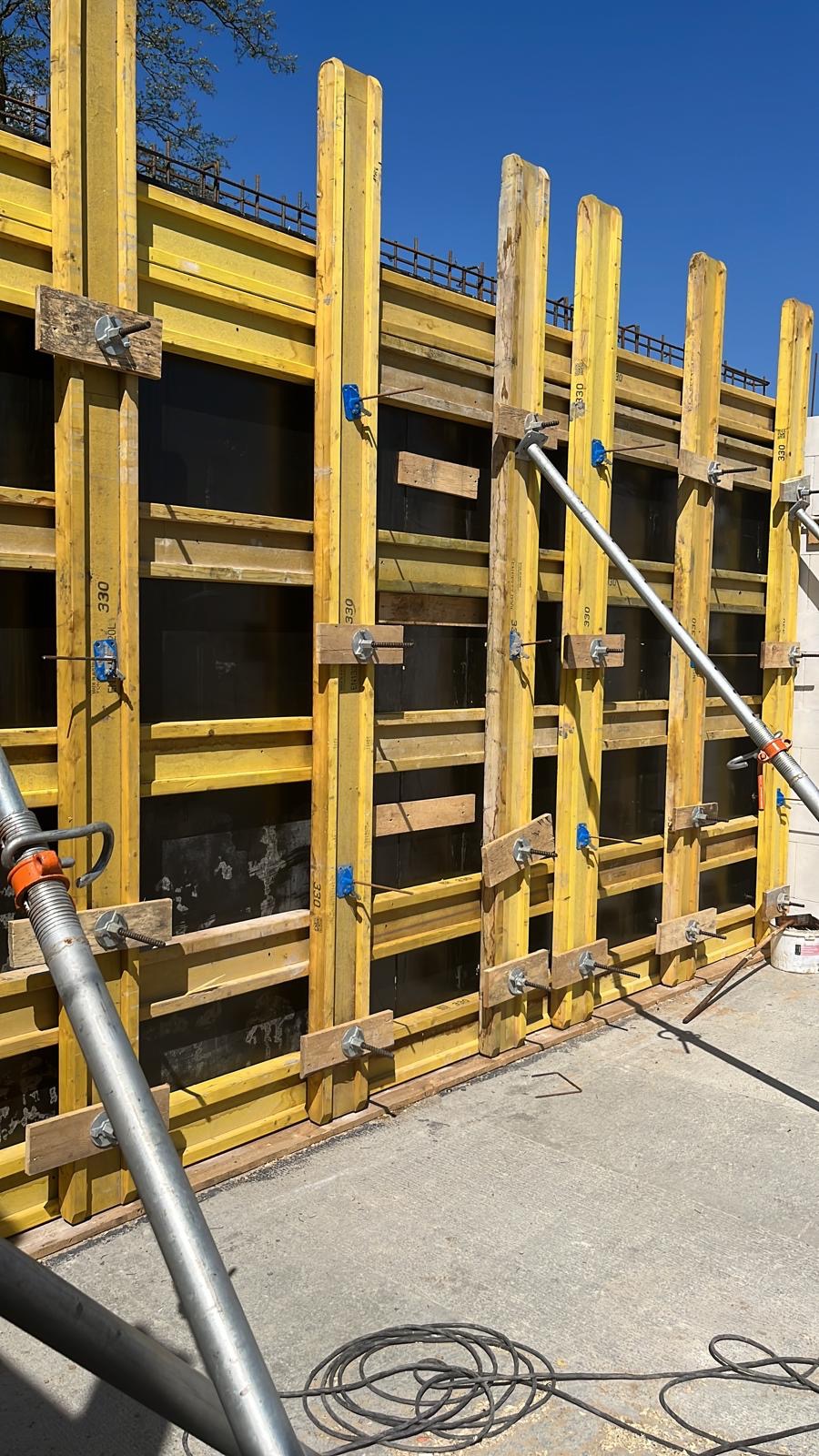 Szczegółowe ujęcie szalunku fundamentowego z żółtymi belkami i stalowymi rozpórkami, przygotowanego do wylewania betonu, widoczne elementy zbrojenia.