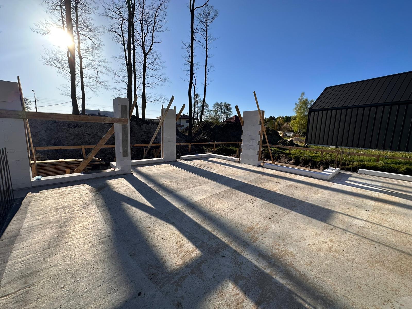Surowy betonowy fundament domu jednorodzinnego w trakcie budowy, widoczne bloczki fundamentowe i drewniane podpory, jasne światło słoneczne rzuca długie cienie na posadzkę.