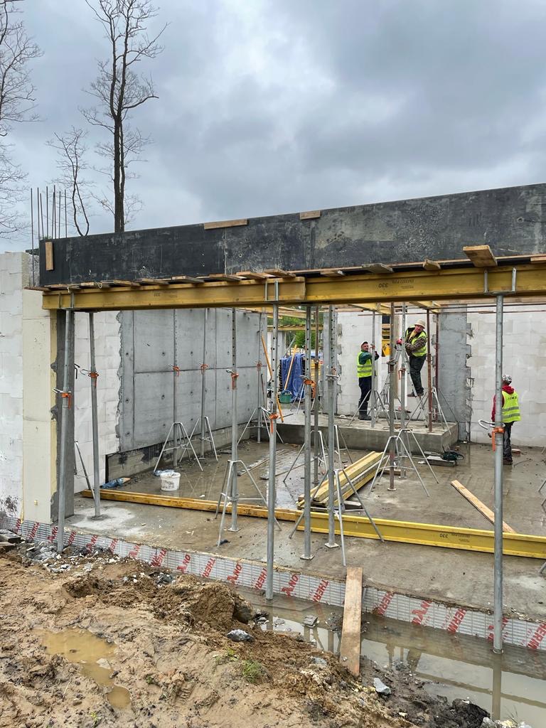 Budowa domu w stanie surowym otwartym, widoczne ściany z bloczków, szalunki stropowe na podporach, oraz pracownicy w odblaskowych kamizelkach na placu budowy w pochmurny dzień.