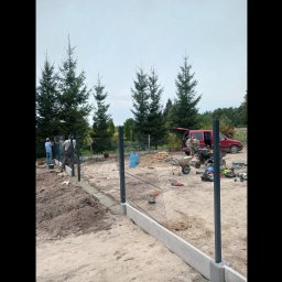 Skotbud - Ekipa montuje metalową siatkę ogrodzeniową na betonowej podmurówce, widoczny samochód dostawczy i narzędzia na placu budowy, w tle wysokie drzewa iglaste.