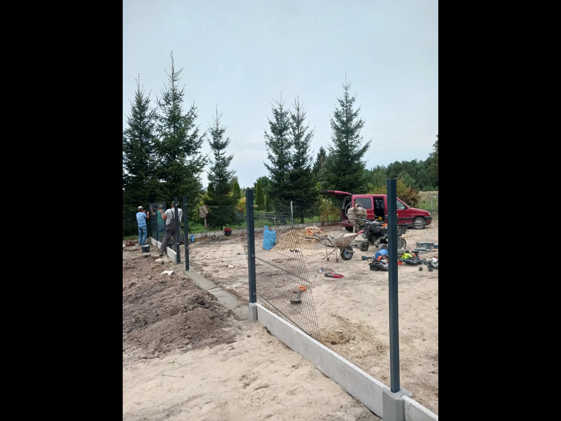 Ekipa montuje metalową siatkę ogrodzeniową na betonowej podmurówce, widoczny samochód dostawczy i narzędzia na placu budowy, w tle wysokie drzewa iglaste.