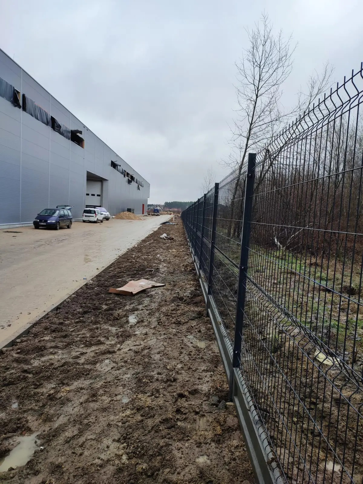 Nowo postawione, czarne, panelowe ogrodzenie na betonowej podmurówce, oddzielające teren przemysłowy z halą magazynową od zarośli, w błotnistym terenie po deszczu.