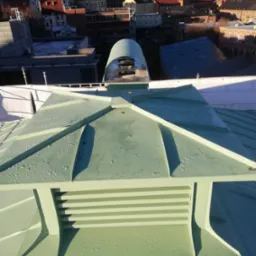 Widok z góry na fragment dachu z blachy w kolorze jasnej zieleni, z widocznymi kroplami wody, na tle zabudowy miejskiej w słoneczny dzień.