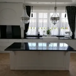 Elegancka, biała kuchnia z wyspą i czarnym blatem, ozdobiona kryształowymi lampami i wzorzystymi zasłonami, widok na okno z białymi firankami.
