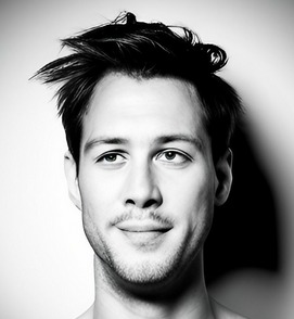 Portret młodego mężczyzny z rozwichrzonymi włosami, uśmiechającego się lekko, w czerni i bieli, oświetlonego z góry.