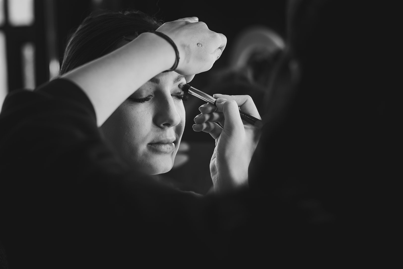 Czarno-białe zbliżenie: aplikacja cienia do powiek pędzlem na powiece kobiety, z zamkniętymi oczami i delikatnym uśmiechem, widoczne krople wody na dłoni wizażysty.