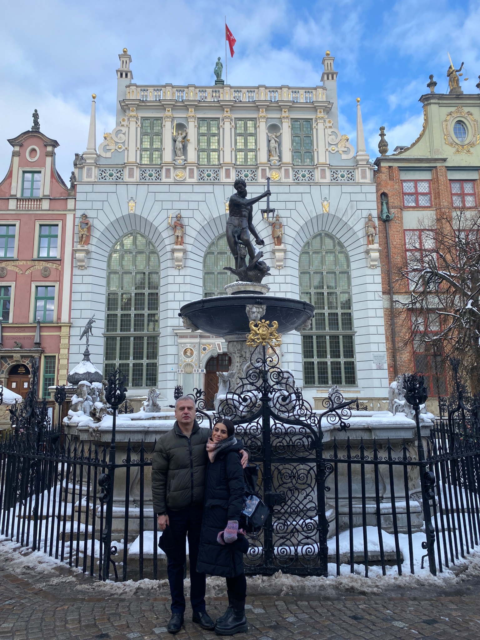 Para ubrana w zimowe kurtki pozuje do zdjęcia na tle Fontanny Neptuna w Gdańsku, w zimowej scenerii.