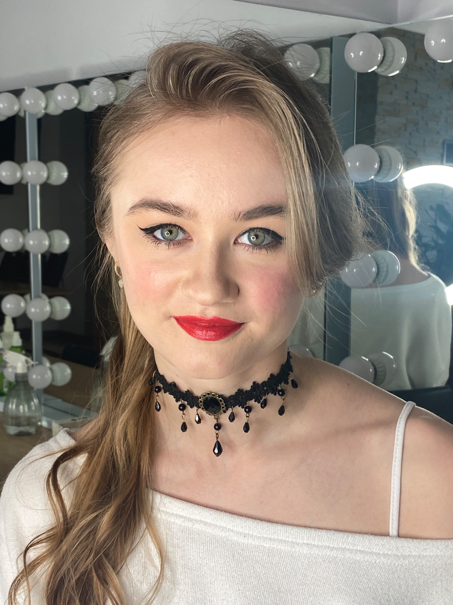 Portret kobiety z czerwonymi ustami, delikatnym makijażem oczu i chokerem z czarnymi koralikami, stojącej przed lustrem z oświetleniem w stylu hollywoodzkim.