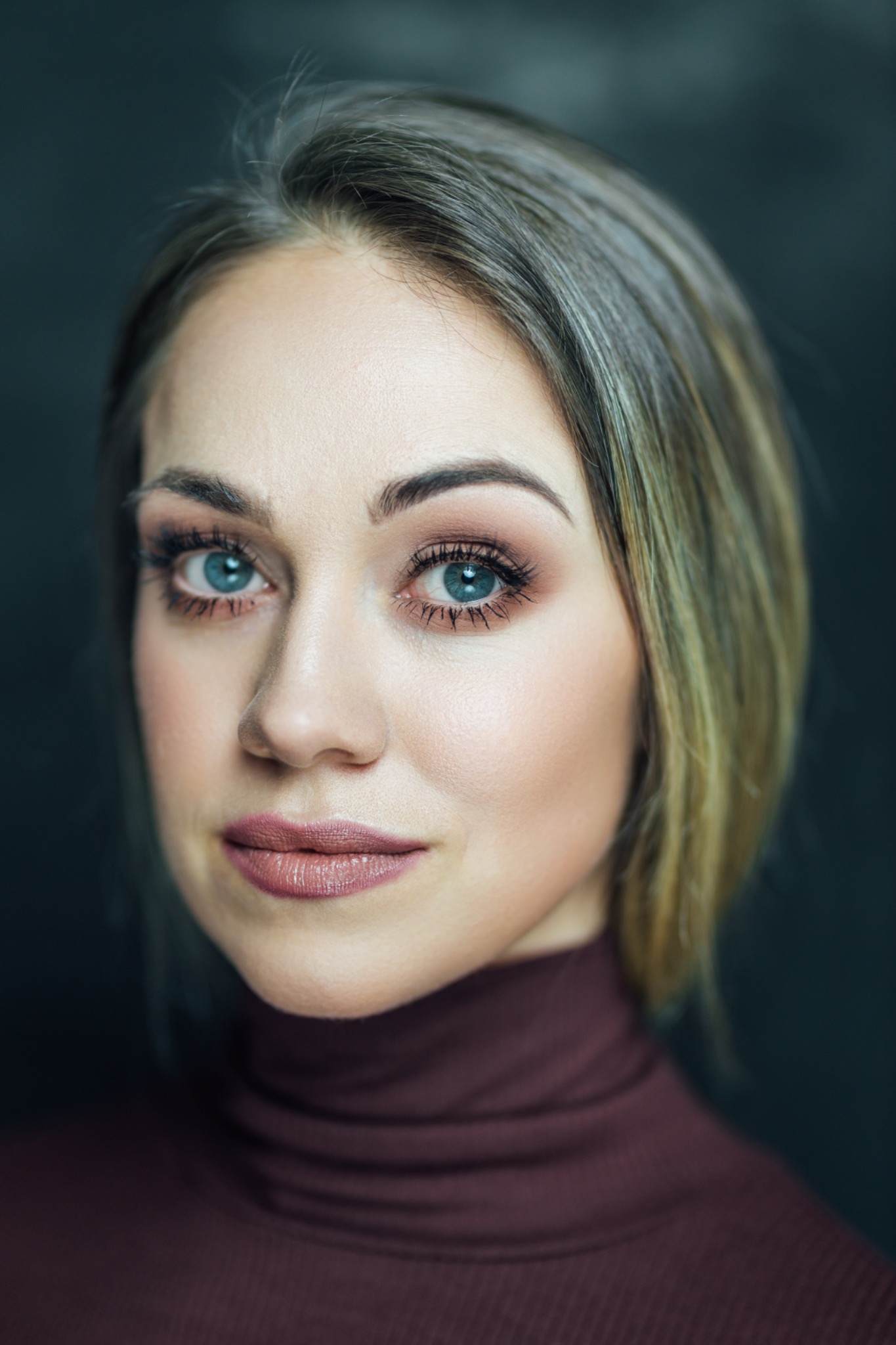 Portret kobiety z delikatnym makijażem, podkreślone oczy i usta w odcieniach różu, włosy blond z odrostami, ciemne tło.