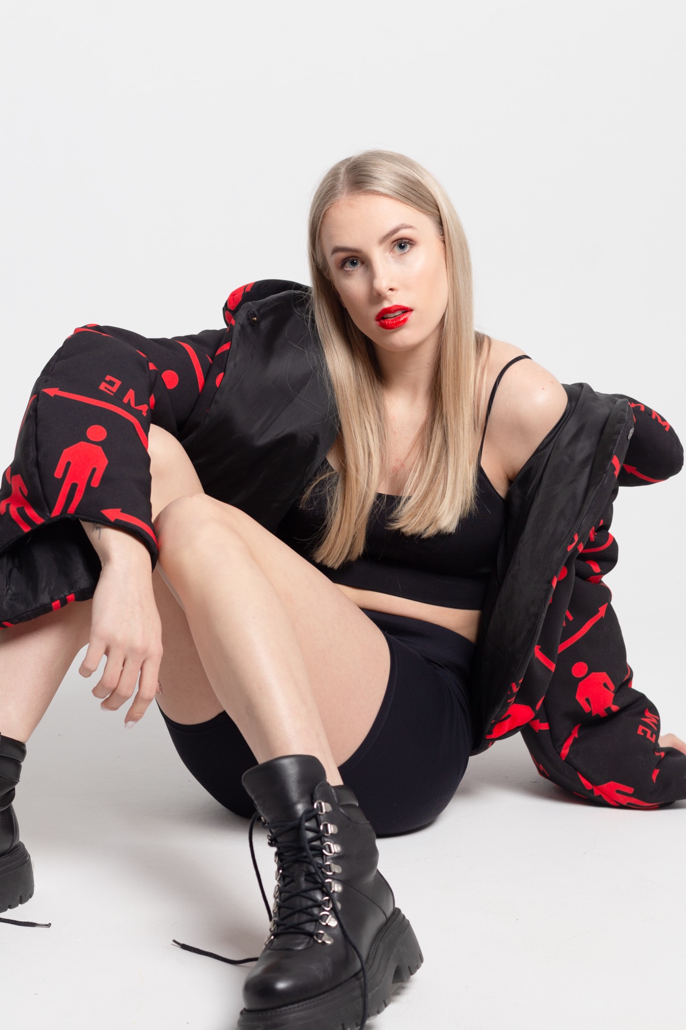 Blondwłosa kobieta z intensywnie czerwoną szminką, pozująca w czarnym topie, spodenkach i ciężkich butach, z nonszalancko zarzuconą, wzorzystą kurtką.