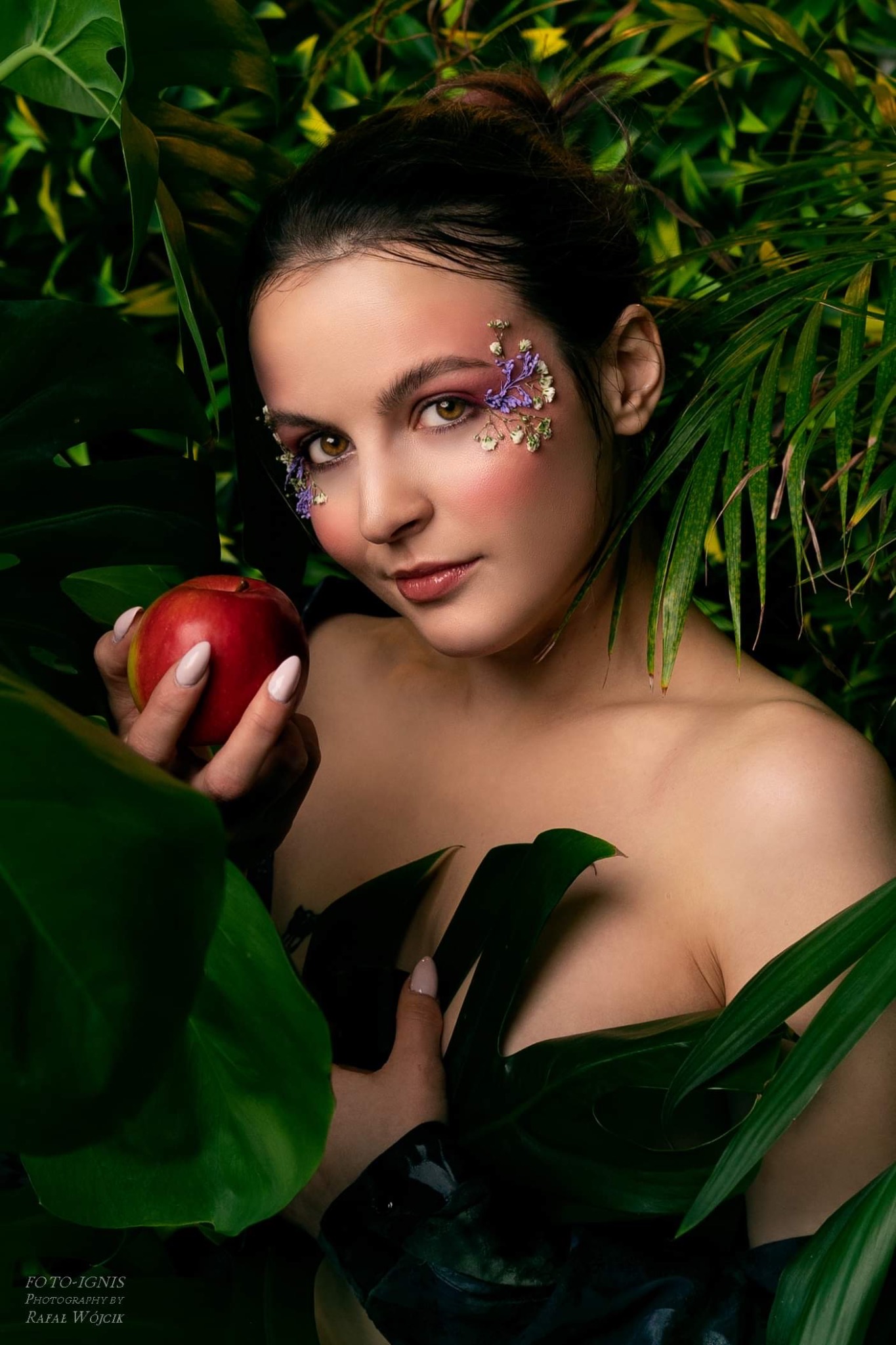 Portret kobiety z kwiatami wokół oczu, trzymającej czerwone jabłko na tle tropikalnej roślinności.