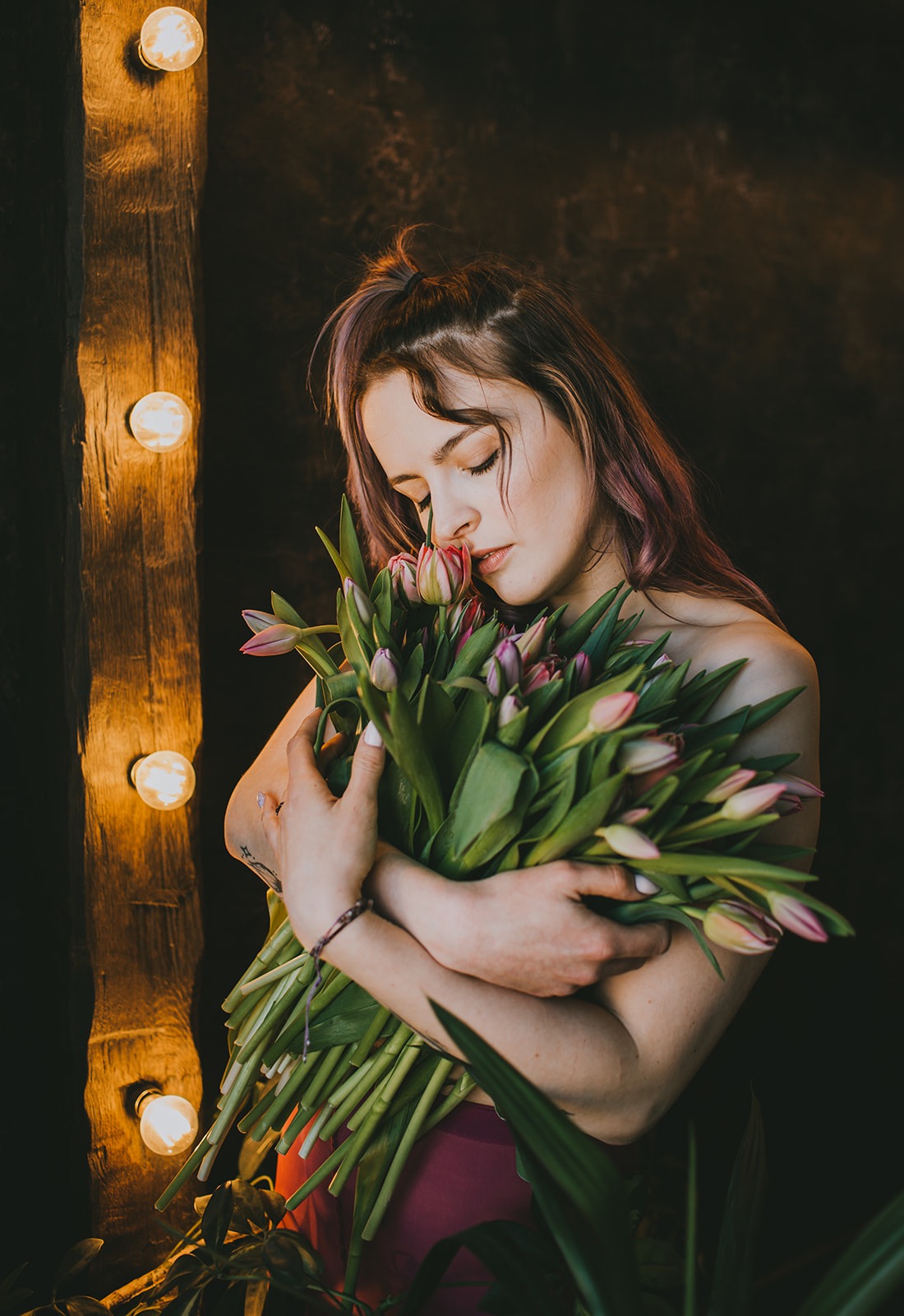 Kobieta z fioletowymi pasemkami, z zamkniętymi oczami, przytula bukiet tulipanów na tle rustykalnej ściany z żarówkami.