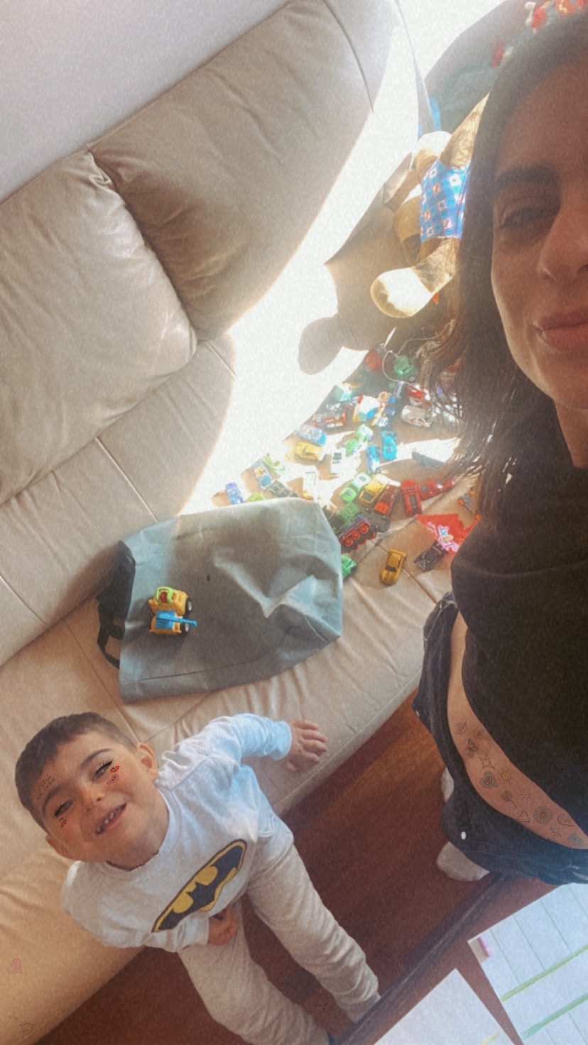 Zdjęcie z góry przedstawia kobietę robiącą selfie z synem, który ma naklejki na twarzy i koszulkę z Batmanem, siedzących na beżowej kanapie z rozrzuconymi zabawkami samochodowymi i szarą torbą.