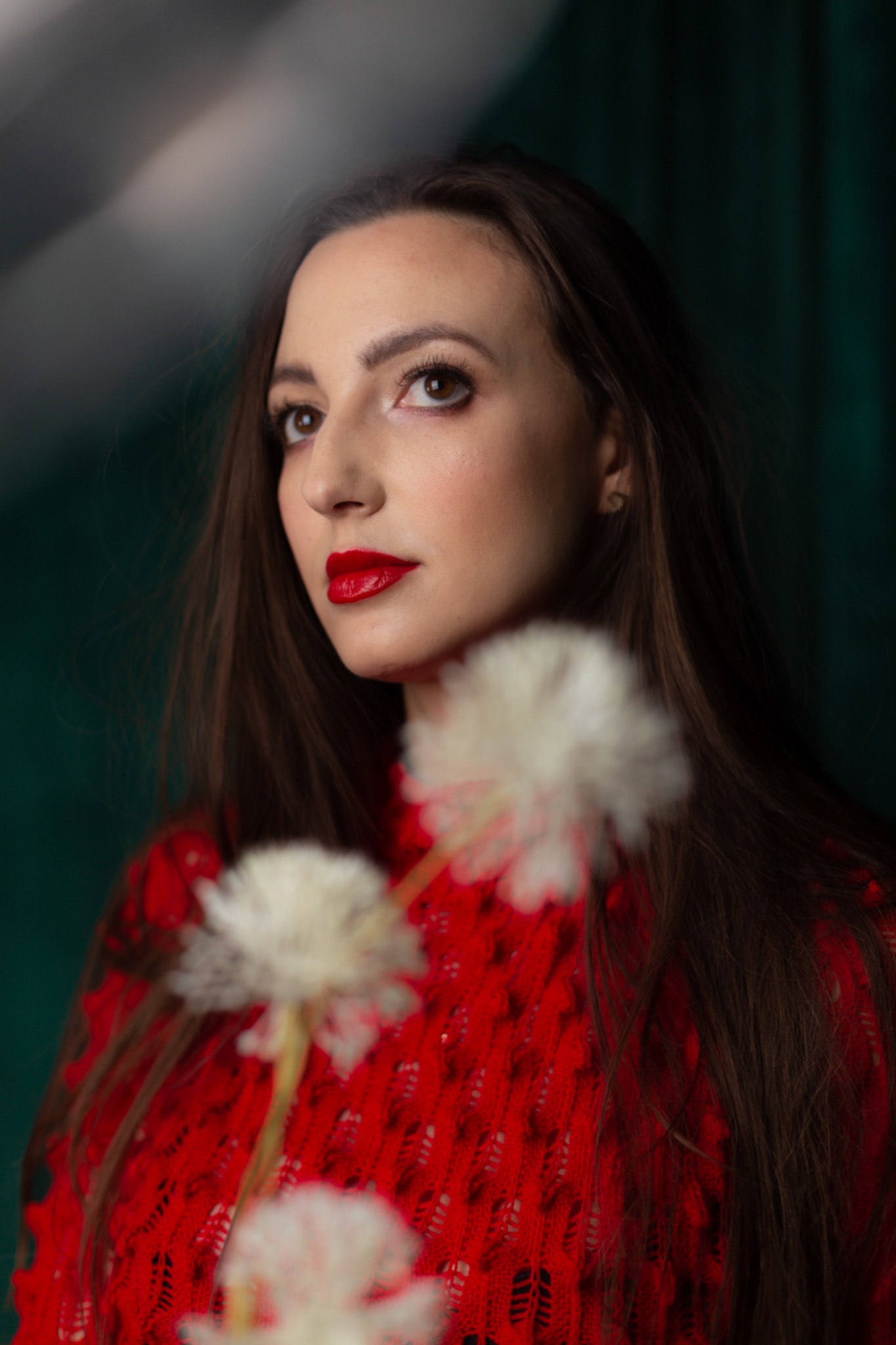Portret kobiety z intensywnie czerwoną pomadką, spoglądającej w górę, z rozmytymi kwiatami na pierwszym planie i ciemnozielonym tłem.