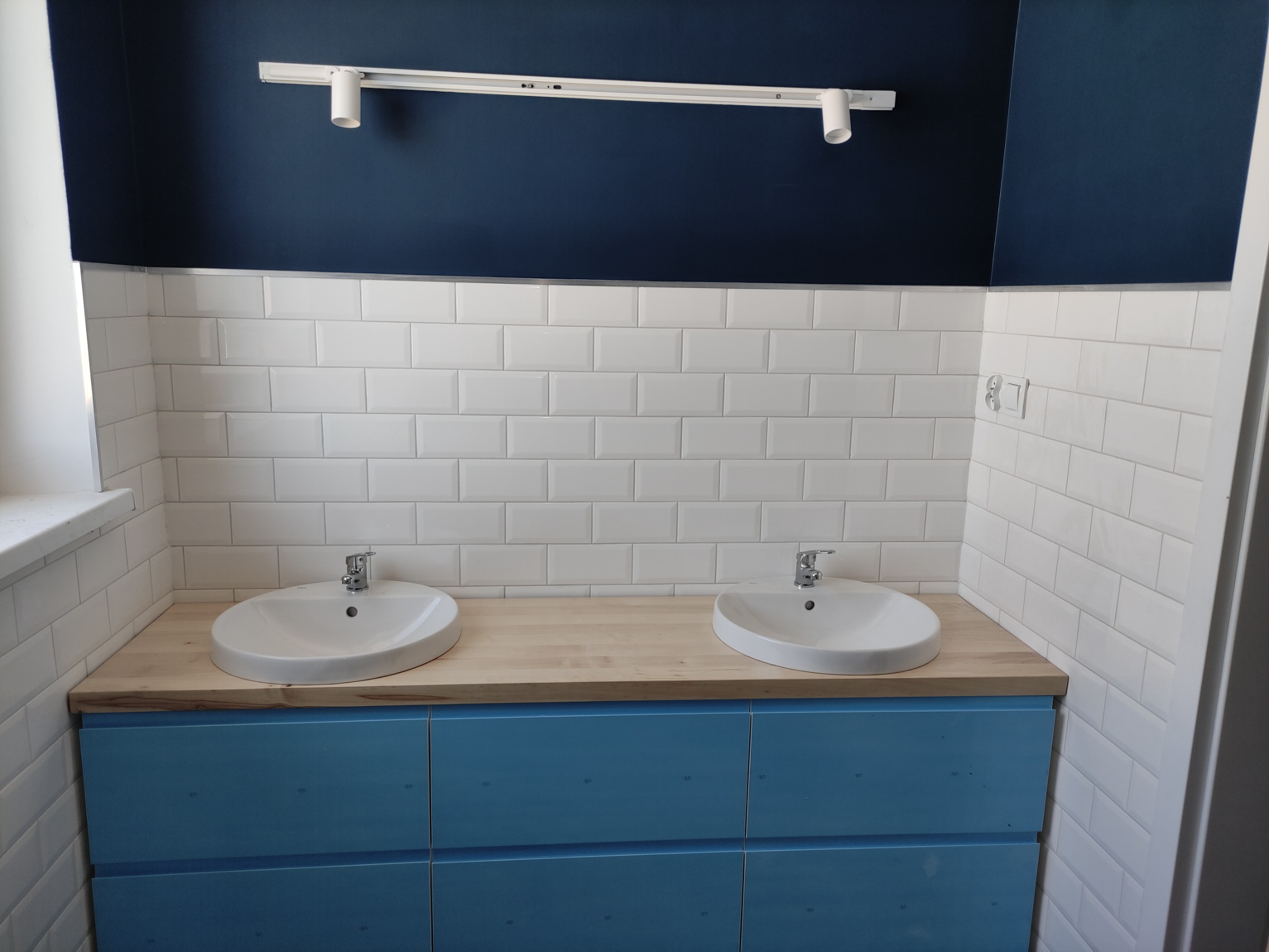 Nowoczesna łazienka z dwoma białymi umywalkami nablatowymi na drewnianym blacie, niebieska szafka pod umywalką, białe kafelki na ścianie i ciemnoniebieski sufit z reflektorami.